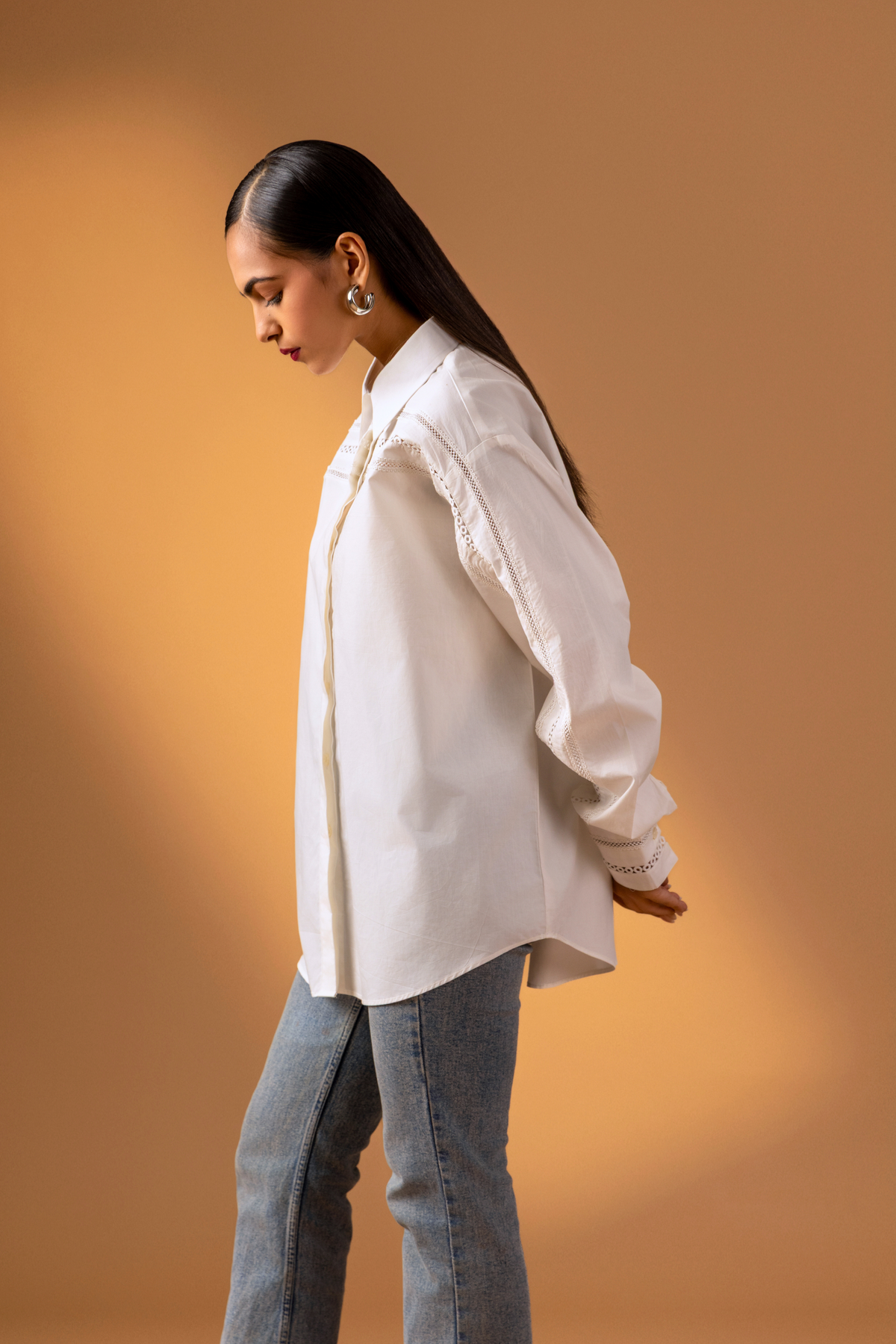 Side view women stands confidently in a stylish white button-up shirt with intricate details and puffed sleeves, paired with blue jeans.