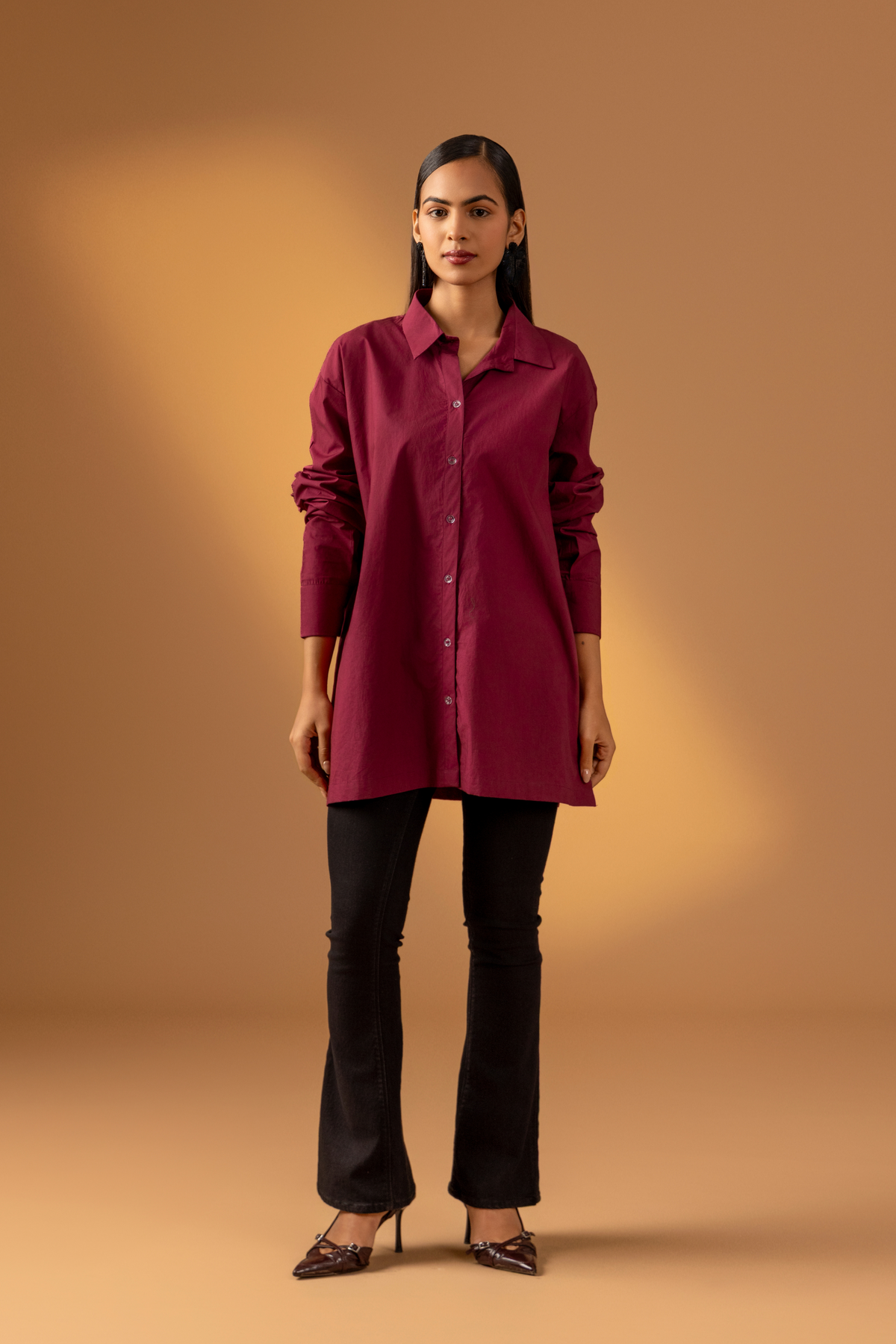 A woman wearing a maroon shirt with long sleeves paired with black pants against a warm, muted background, showcasing a stylish, casual look. 