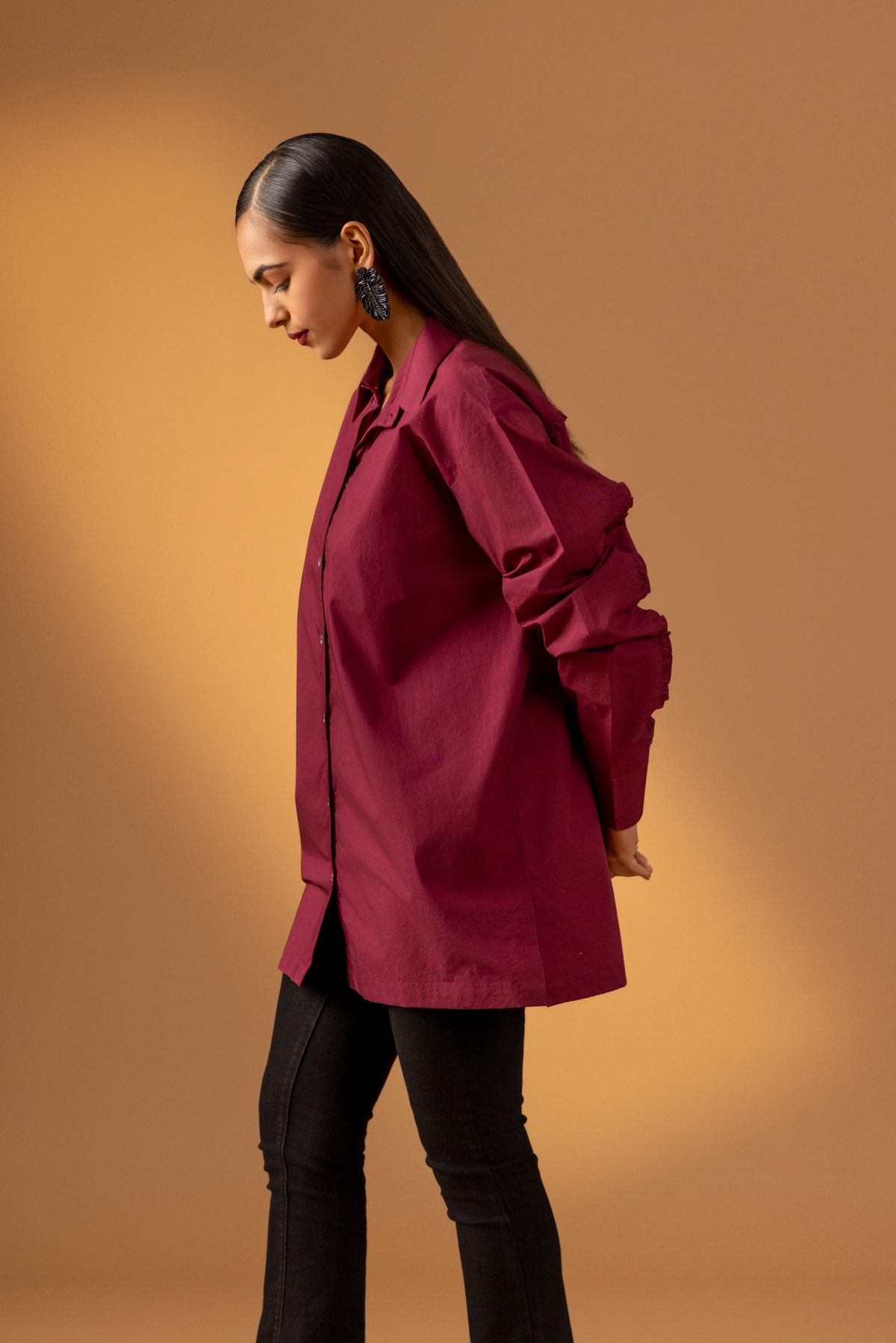 Side view of a woman wearing a maroon shirt with long sleeves paired with black pants against a warm, muted background, showcasing a stylish, casual look. 