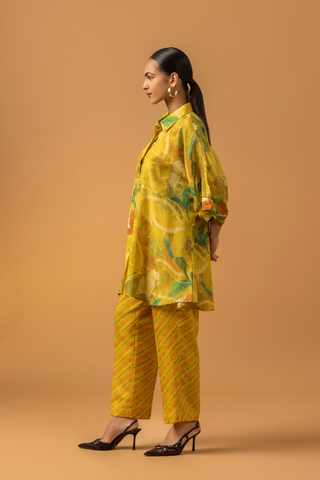 Side view of a model stands in a bright yellow co-ord set featuring a patterned oversized shirt and matching striped pants against a warm backdrop.