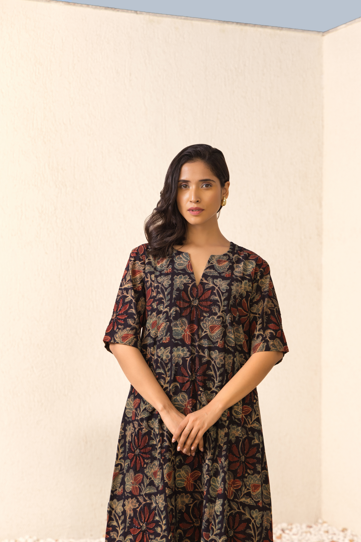 A woman stands confidently in a ajrakh print patterned co-ord set against a neutral wall background, hands clasped in front of her.