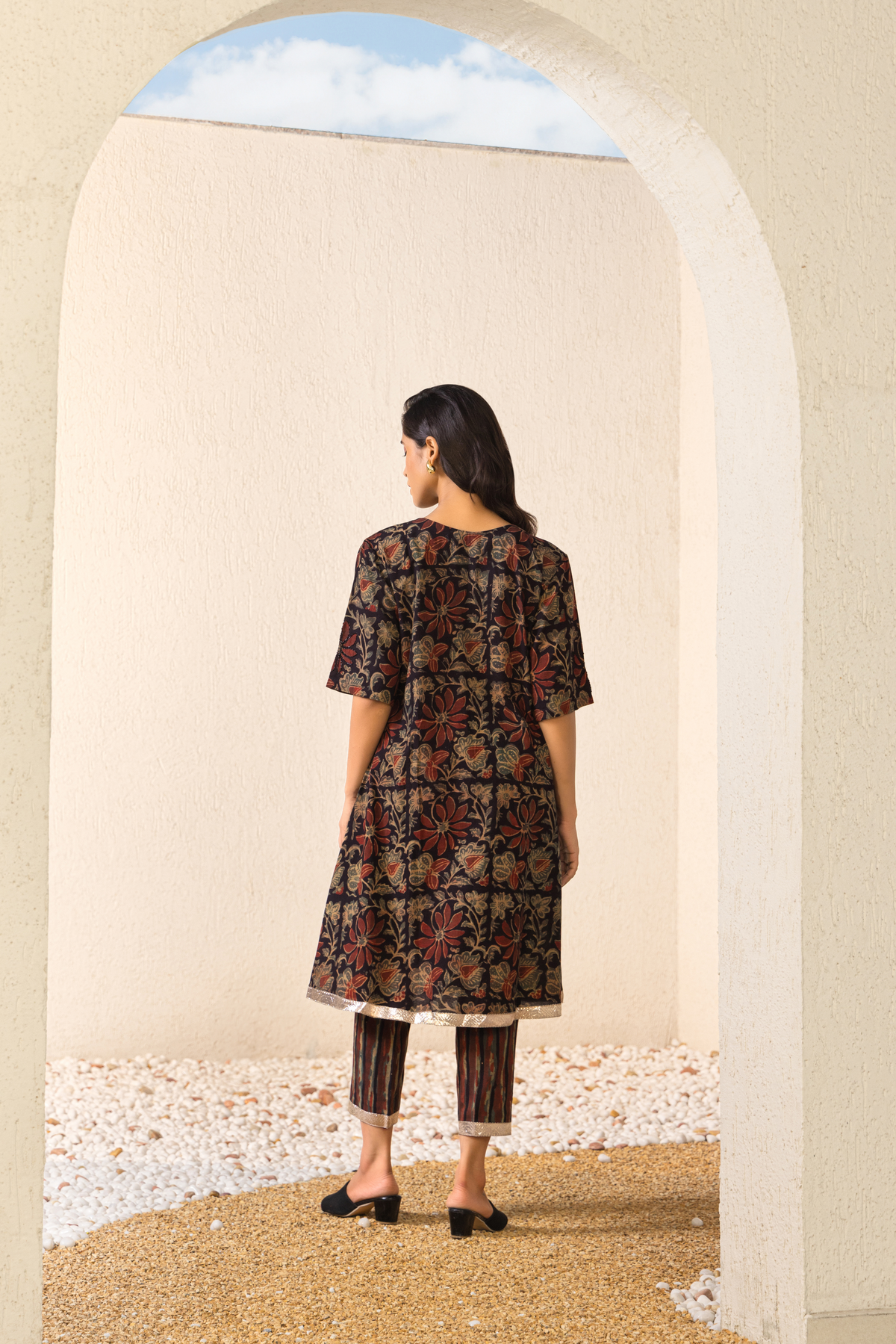 Back View of a woman wearing a dark floral-patterned traditional outfit with a golden hemline, paired with striped pants and black sandals, standing under a light-colored archway on a gravel path.