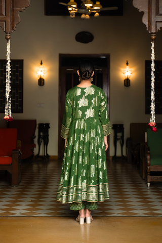 Back view of a woman in a vibrant green anarkali kurta set with matching pants stands gracefully in a well-decorated interior.
