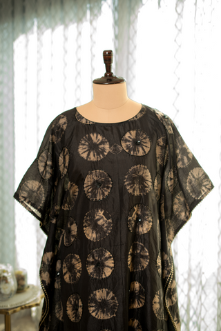 Closeup view of a black kaftan dress with beige circular tie-dye patterns, displayed on a mannequin in a softly lit room.