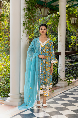 A person wears a vibrant turquoise and yellow handblock printed kurta set, standing gracefully on a checkered floor amidst lush greenery.
