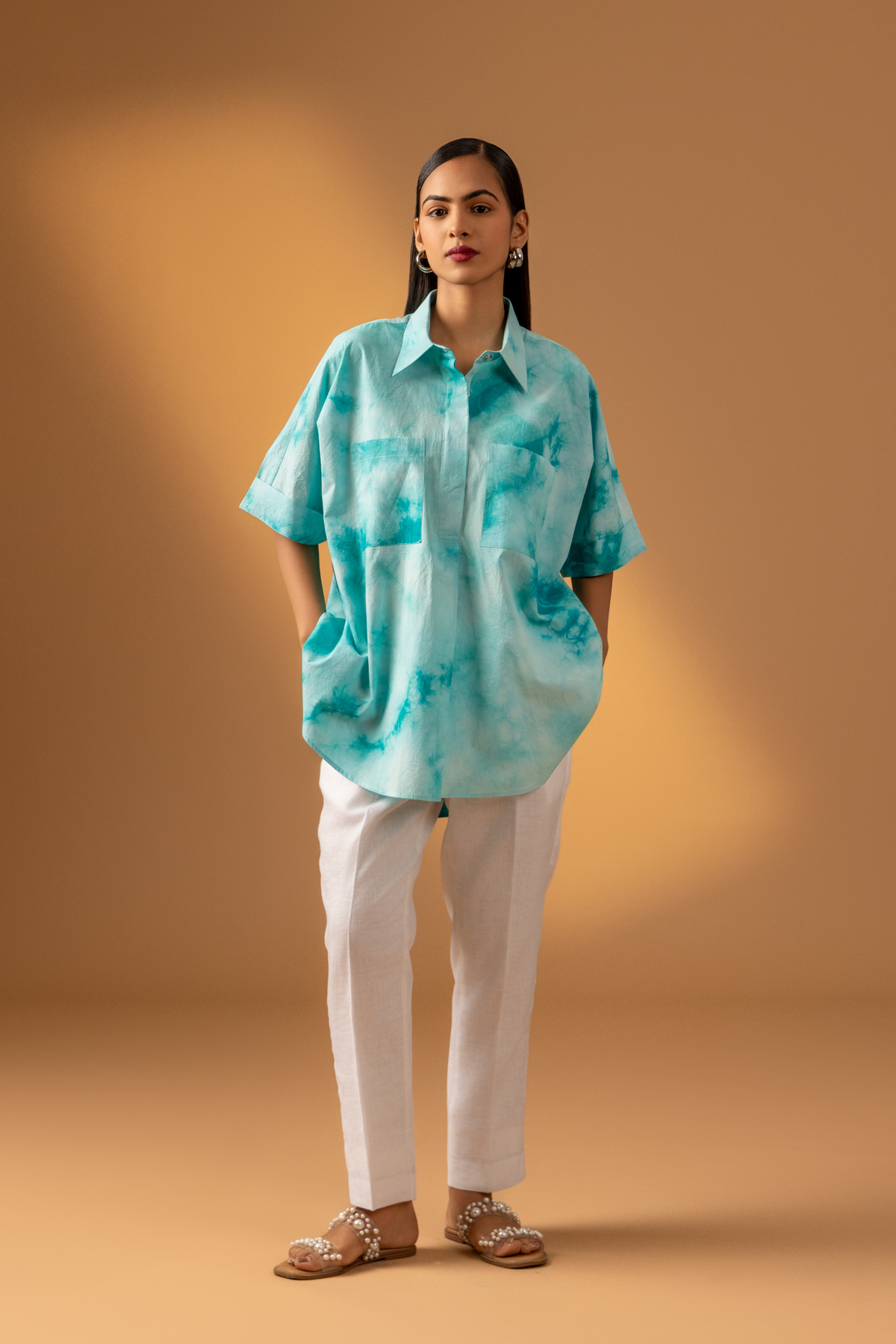 A person stands confidently in a teal tie-dye oversized shirt and light-colored trousers against a warm, neutral background.