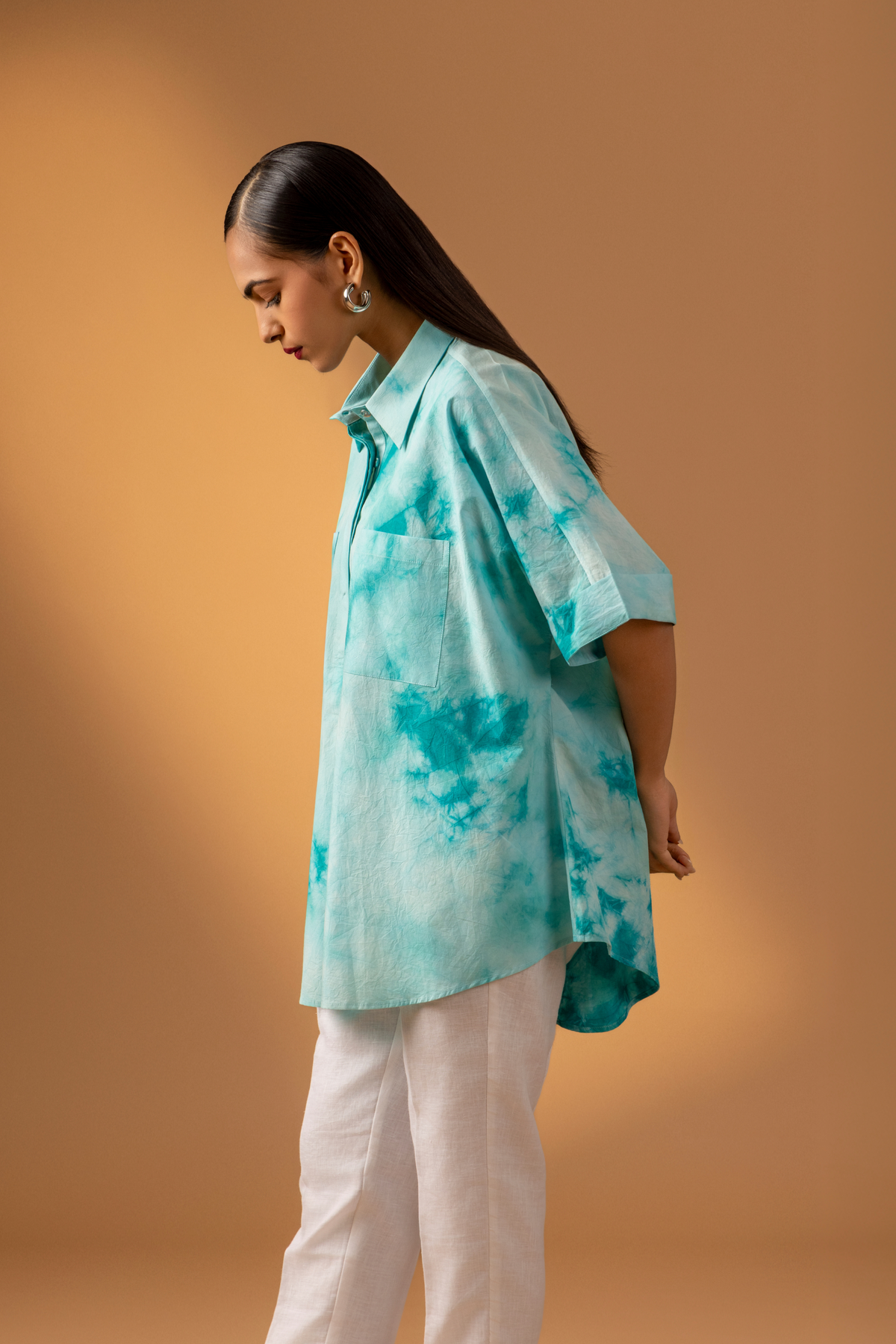 Side view if a woman stands confidently in a teal tie-dye oversized shirt and light-colored trousers against a warm, neutral background.