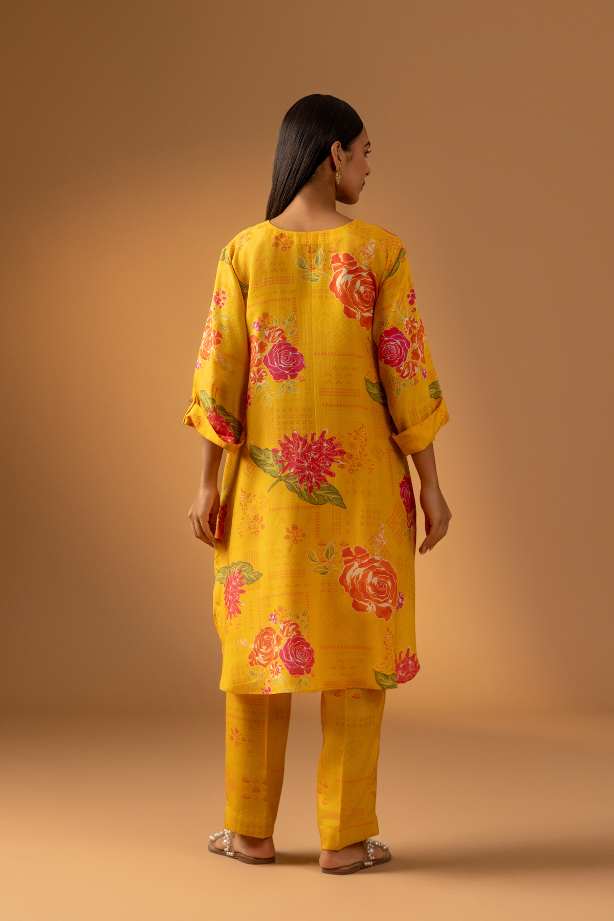 Back view of a woman in a vibrant yellow floral co-ord set and matching pants stands confidently against a warm, neutral background.