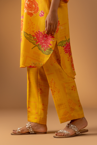 A close-up of a woman wearing a vibrant yellow floral co-ord set and matching pants stands confidently against a warm, neutral background.