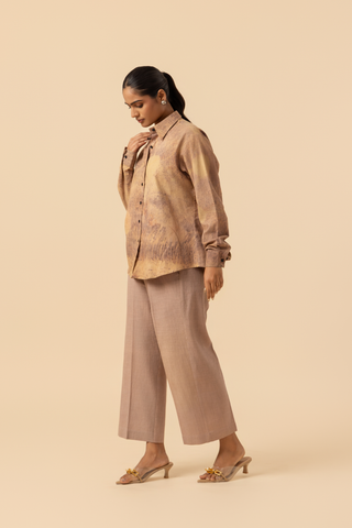 Side view of a model stands against a light beige background, wearing a patterned co-ord set which has button-up shirt and wide-legged beige pants, accessorized with heels.