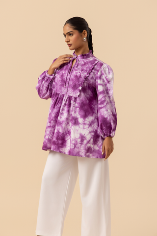 Side view of a model wears a vibrant purple tie-dye shirt dress with puffed sleeves, paired with white wide-leg trousers and stylish sandals.