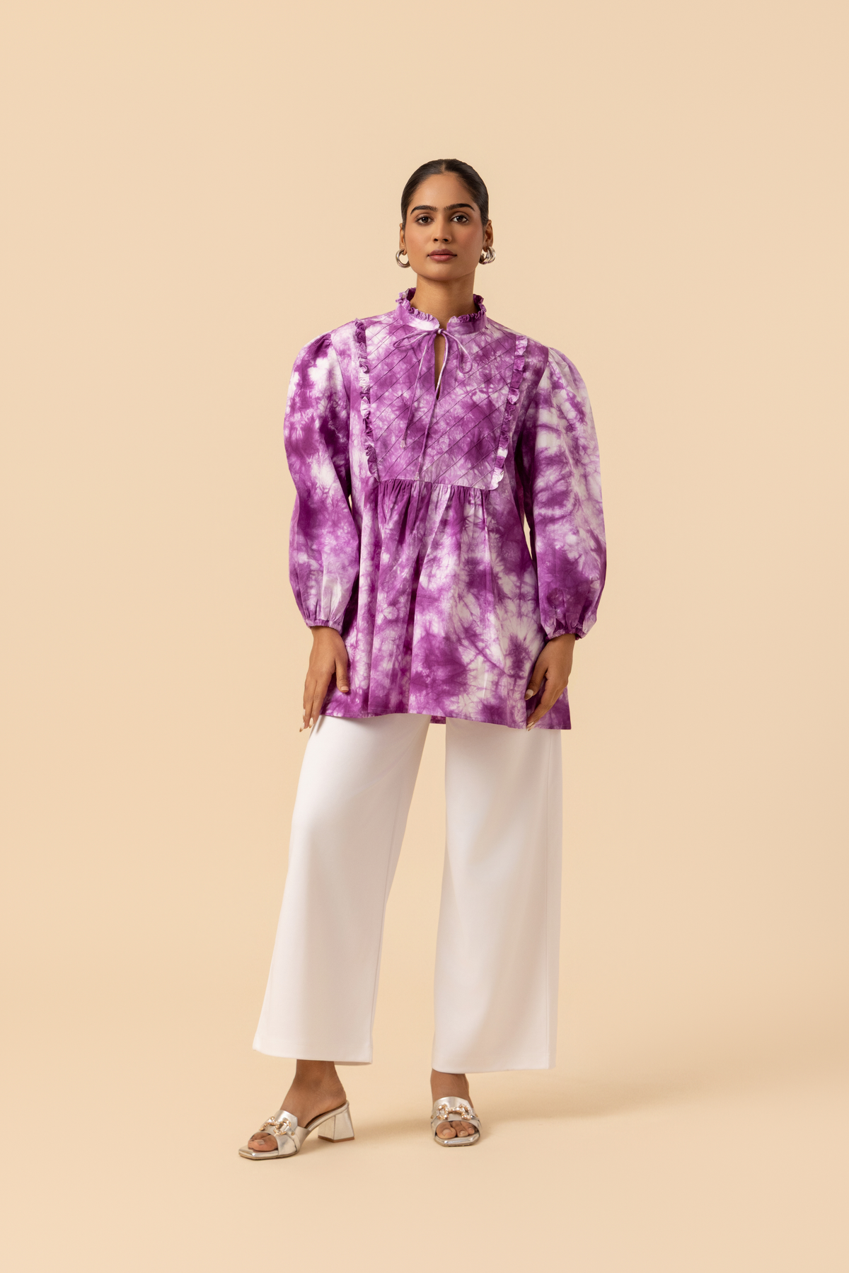 A model wears a vibrant purple tie-dye shirt dress with puffed sleeves, paired with white wide-leg trousers and stylish sandals.