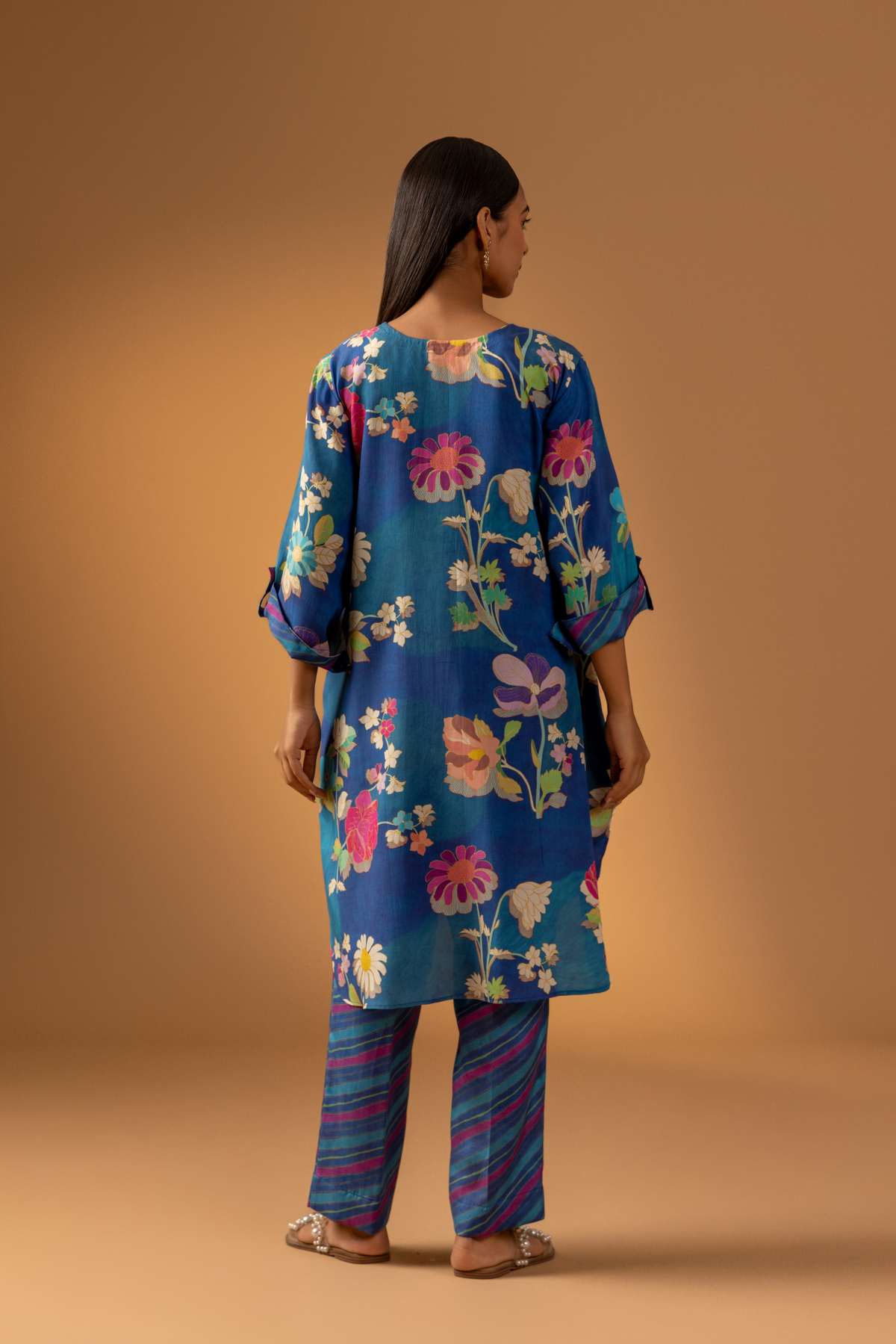 Back view of a woman models a vibrant floral co-ord set paired with striped pants, set against a warm, neutral background.