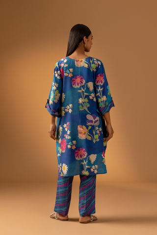 Back view of a woman models a vibrant floral co-ord set paired with striped pants, set against a warm, neutral background.