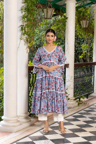 A woman stands gracefully on a checkered marble floor, wearing a floral patterned handblock printed kurta set dress, surrounded by lush greenery and elegant columns.