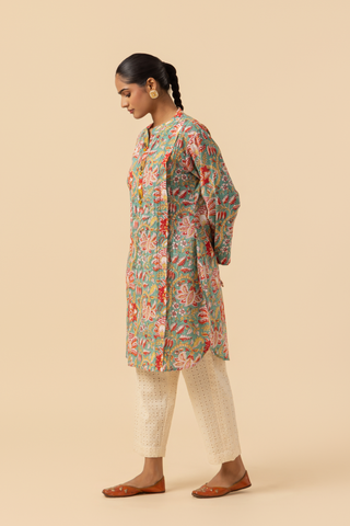 Side view of a person stands elegantly in a colorful floral co-ord set paired with white embellished pants and traditional sandals against a soft beige background.