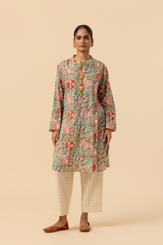 A person stands elegantly in a colorful floral co-ord set paired with white embellished pants and traditional sandals against a soft beige background.