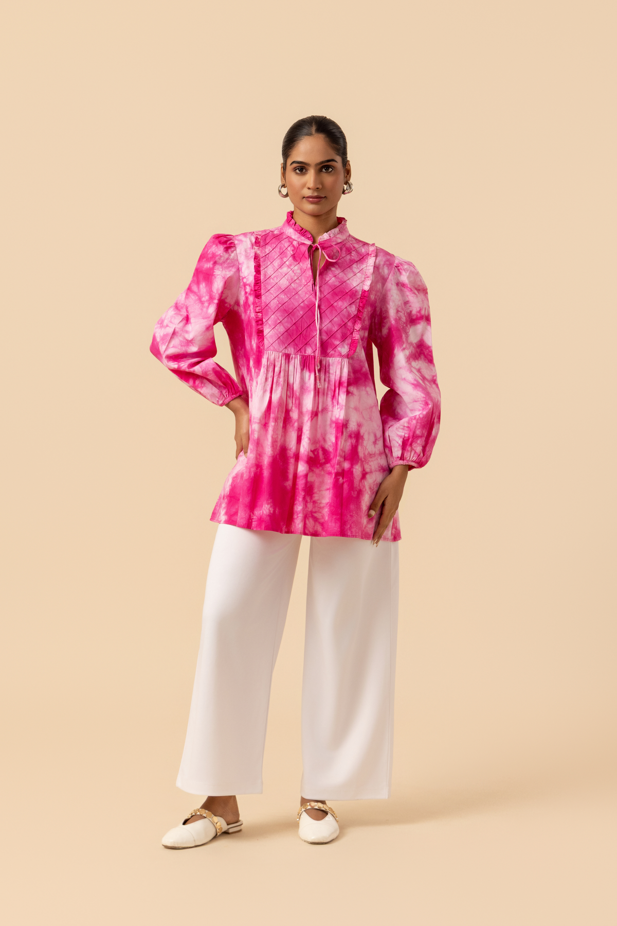 A model wears a vibrant pink tie-dye shirt dress with puffed sleeves, paired with white wide-leg trousers and stylish sandals.