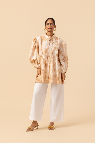 A model wears a vibrant caramel tie-dye shirt dress with puffed sleeves, paired with white wide-leg trousers and stylish sandals.