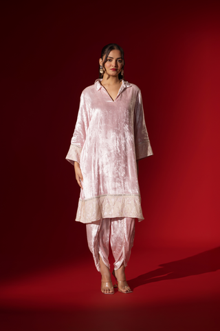 A woman stands in a reflective pink velvet co-ord set with a long tunic and draped pants, against a vibrant red backdrop.