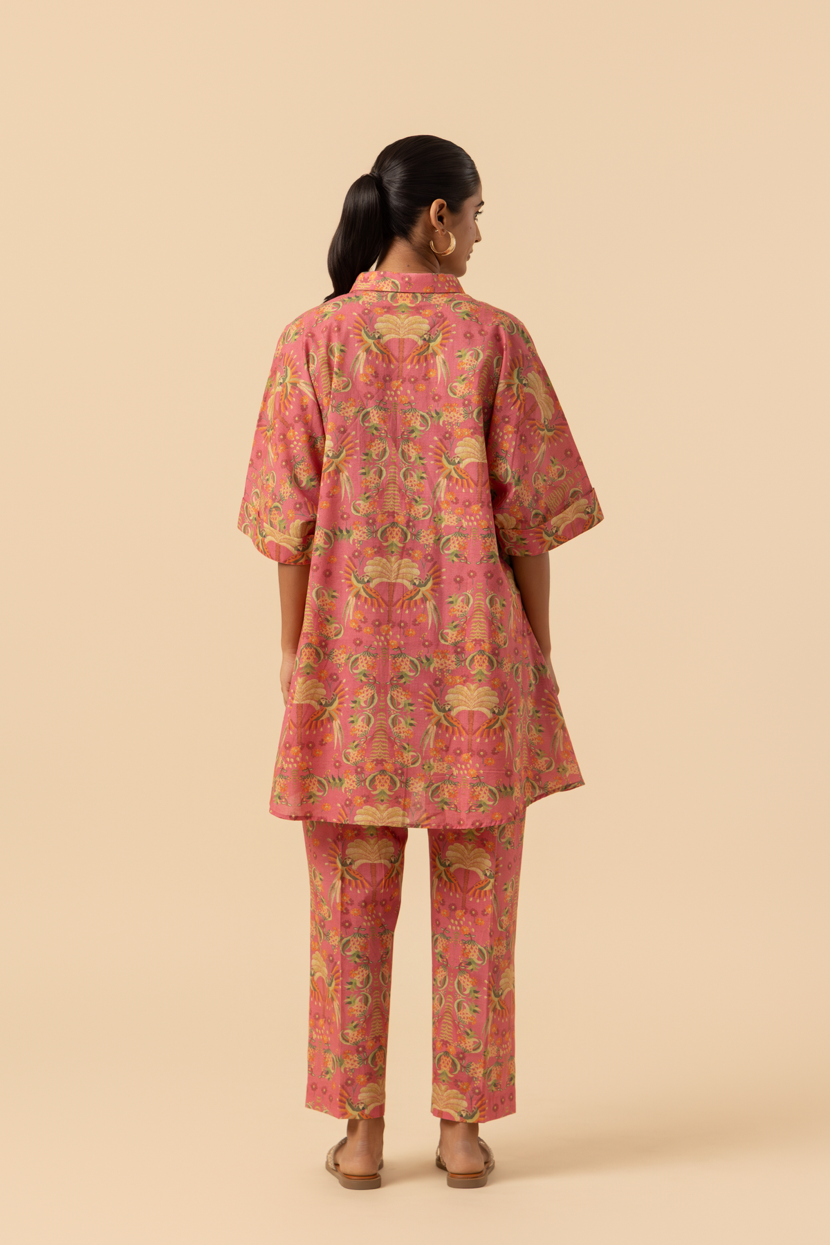 Back view of a woman stands in a vibrant pink and floral-patterned co-ord set, featuring a loose shirt and matching pants, set against a neutral background.