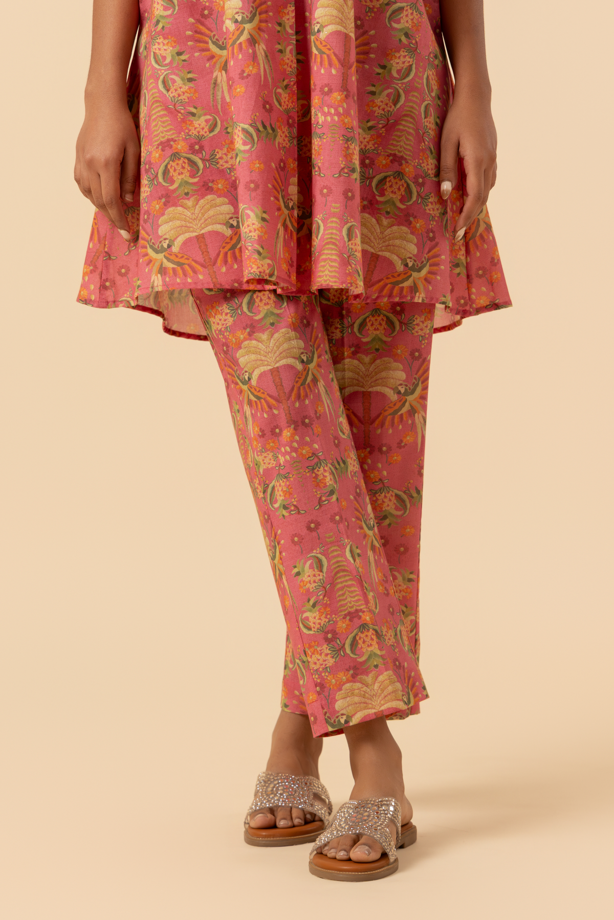 A person stands in a vibrant pink and floral-patterned co-ord set, featuring a loose shirt and matching pants, set against a soft beige background against a neutral backdrop.