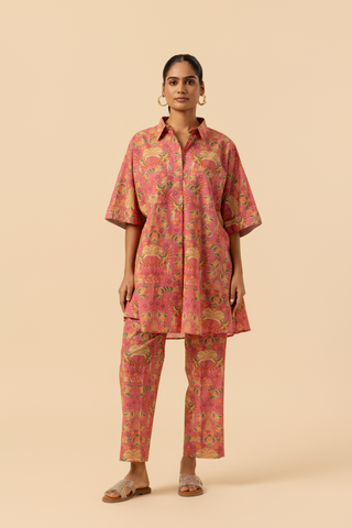 A woman stands in a vibrant pink and floral-patterned co-ord set, featuring a loose shirt and matching pants, set against a neutral background.