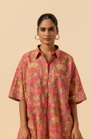 Closeup view of a woman stands in a vibrant pink and floral-patterned co-ord set, featuring a loose shirt and matching pants, set against a neutral background.