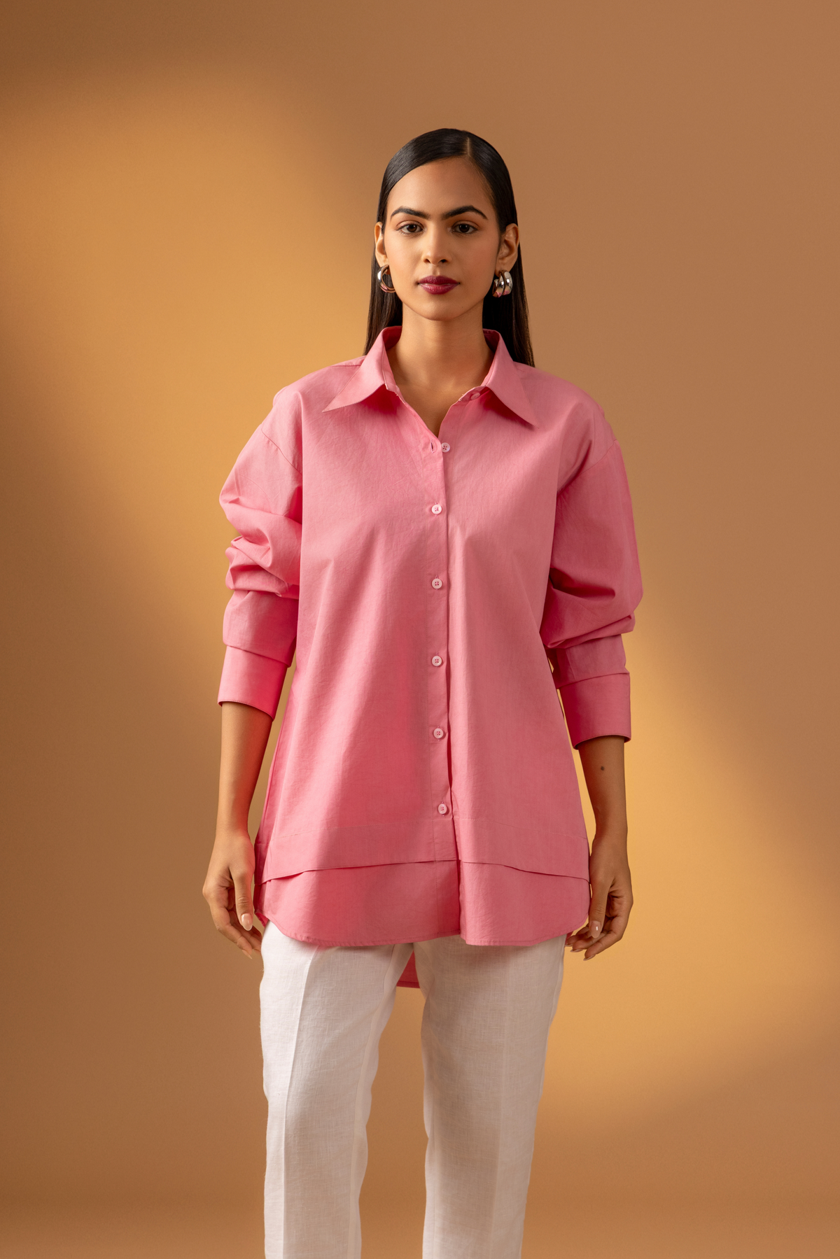 A Woman wearing a pink shirt paired with white trousers, and sandals against a warm, neutral background.