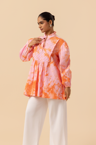 Side view of a model wears a vibrant light pink & orange tie-dye shirt dress with puffed sleeves, paired with white wide-leg trousers and stylish sandals.
