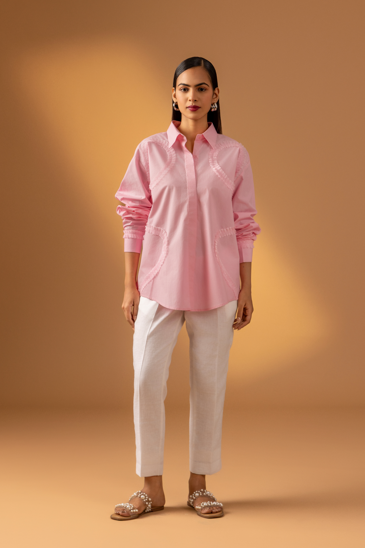 A woman stands against a warm, neutral backdrop, wearing a pink oversized shirt, white trousers, and stylish sandals.