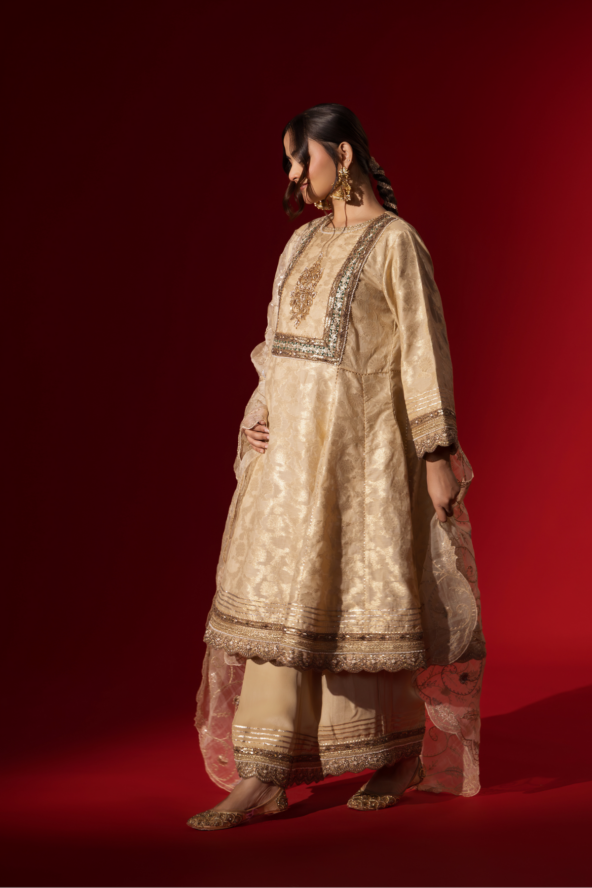 Side view of a woman wearing an elegant cream embroidered sequin brocade kurta set with gold accents and a sheer dupatta, set against a rich red background