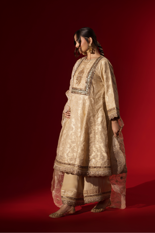 Side view of a woman wearing an elegant cream embroidered sequin brocade kurta set with gold accents and a sheer dupatta, set against a rich red background