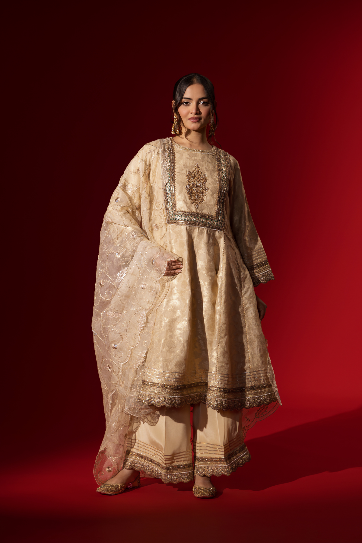 A woman wearing an elegant cream embroidered sequin brocade kurta set with gold accents and a sheer dupatta, set against a rich red background