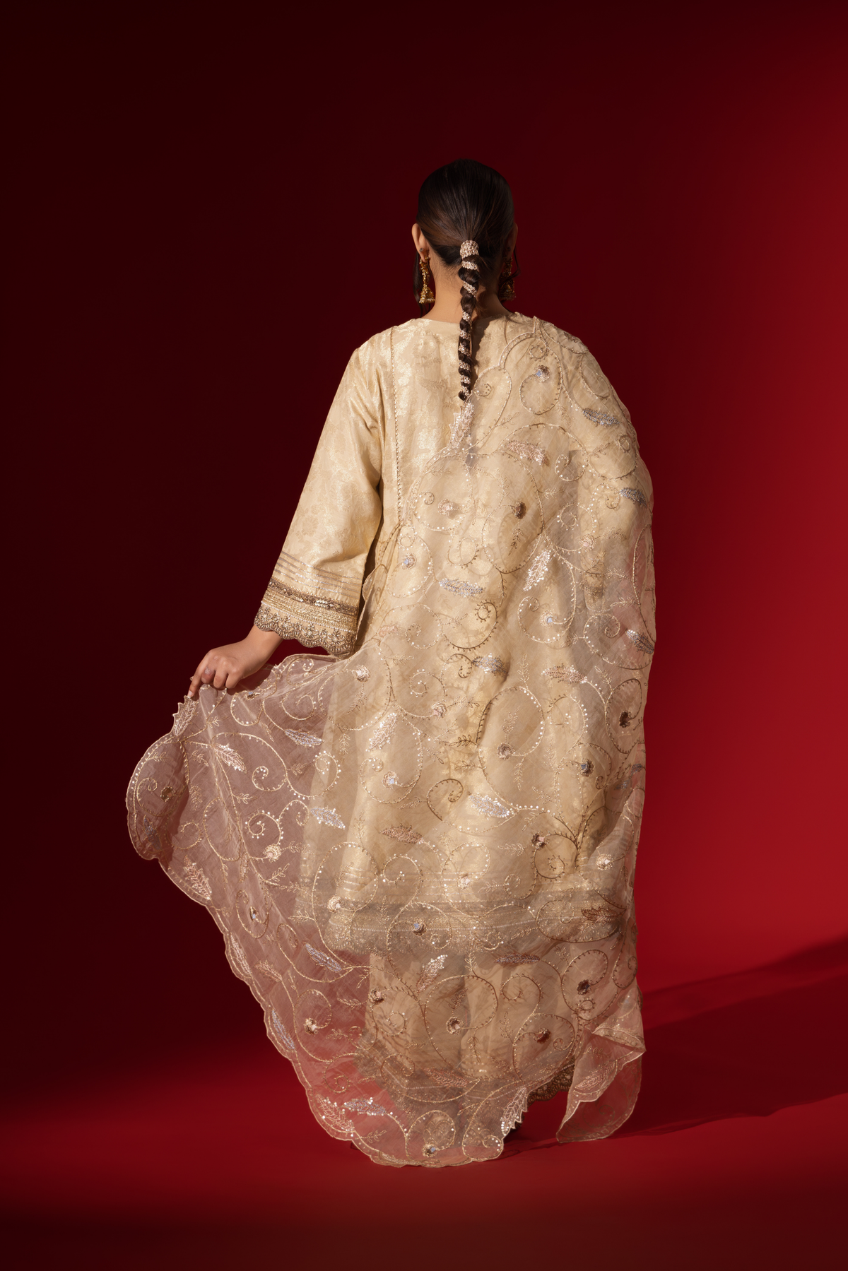 Back view of a woman wearing an elegant cream embroidered sequin brocade kurta set with gold accents and a sheer dupatta, set against a rich red background