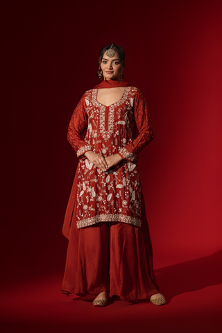 A woman poses in an elegant red embroidered kurta set adorned with intricate white embroidery against a rich red background.