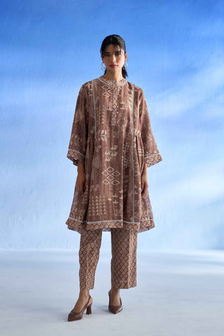 A woman modeling a brown and beige patterned linen co-ord set with matching pants and brown heels, standing against a blue studio backdrop.