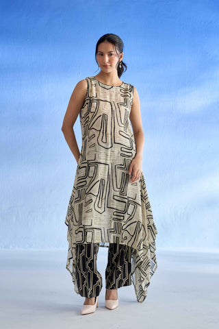 A woman modeling a sleeveless high-low linen co-ord set with an abstract black and beige geometric pattern, paired with black pants featuring beige lines, set against a blue gradient studio background.