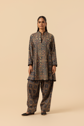 A model wearing a long, patterned ajrakh print co-ord set which has tunic and loose pants, showcasing intricate designs in dark blues and golds against a neutral background.