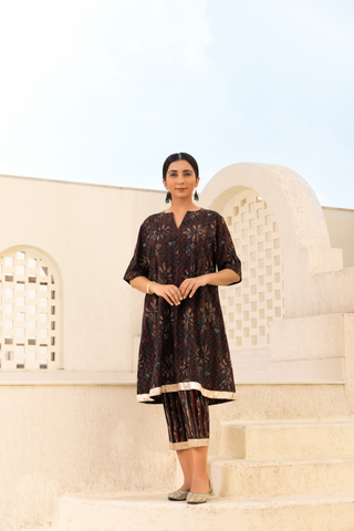 Model dressed in a brown printed kurta set with intricate patterns, paired with striped trousers, styled in a serene outdoor setting