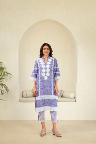 A woman wearing a purple traditional outfit with intricate white embroidery and lace detailing, paired with matching pants, standing in front of an arched alcove in a neutral-toned interior.