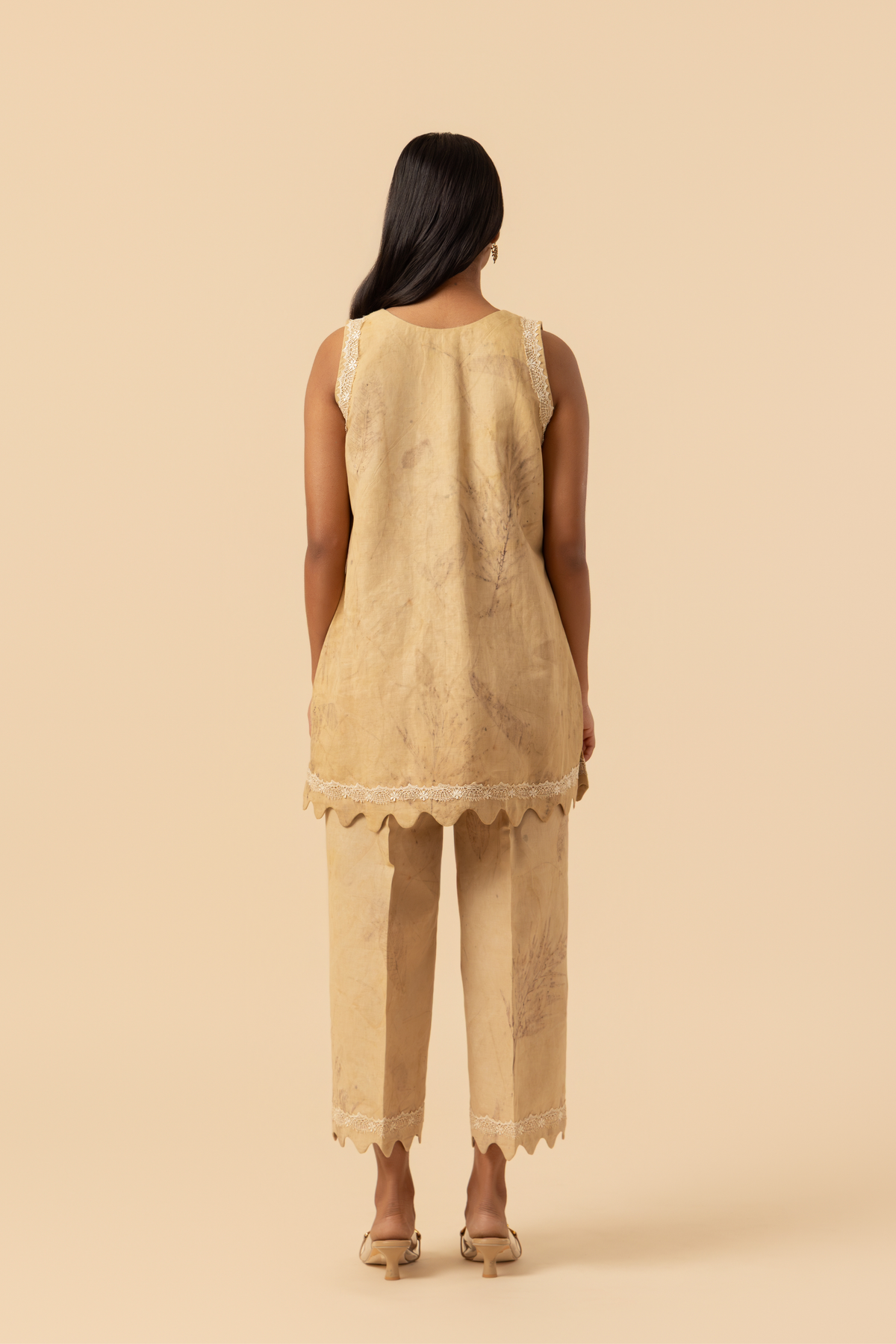 Back view of a person stands in a light beige outfit featuring a sleeveless top and matching pants, complemented by delicate lace detailing.