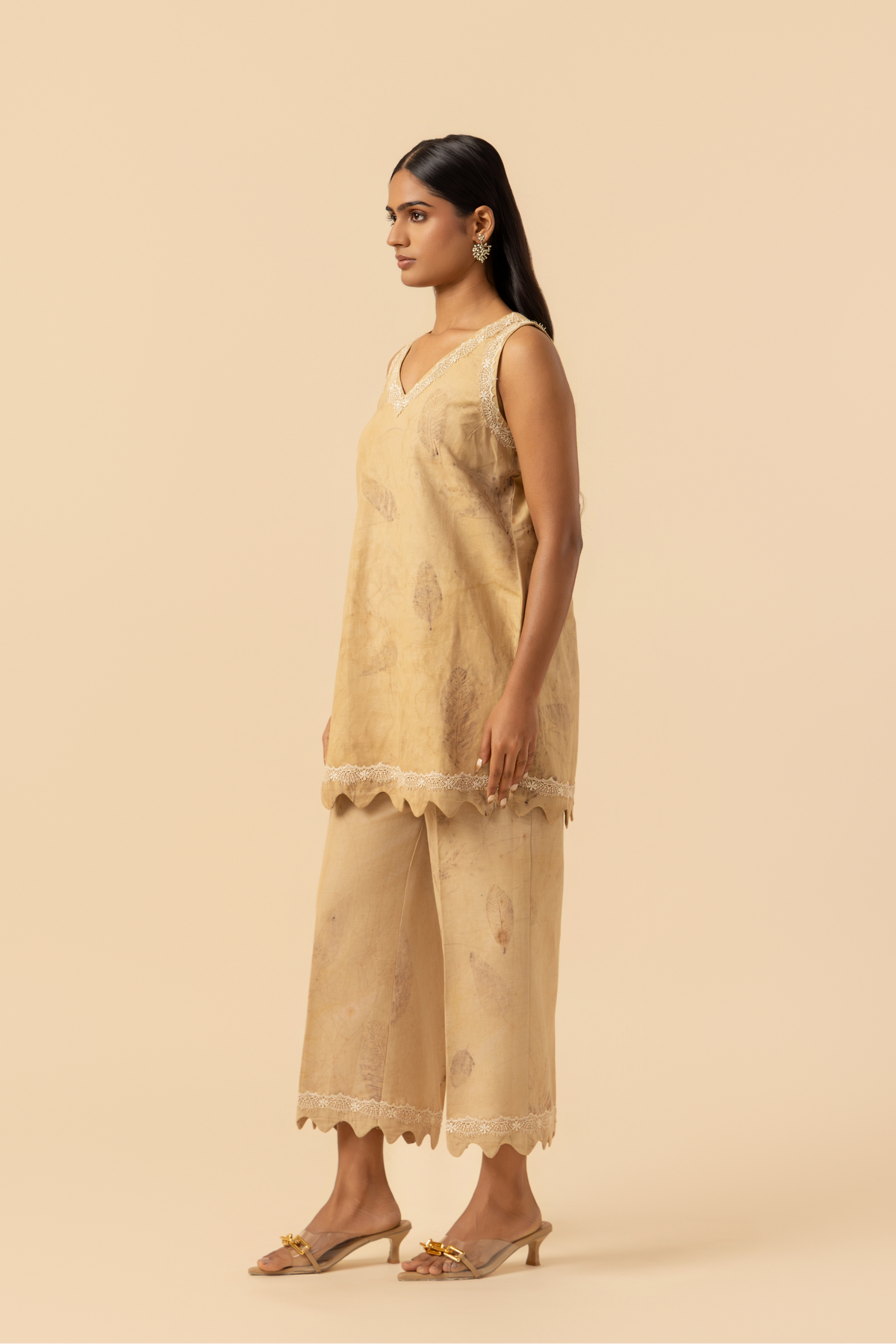 Side view of a person stands in a light beige outfit featuring a sleeveless top and matching pants, complemented by delicate lace detailing.