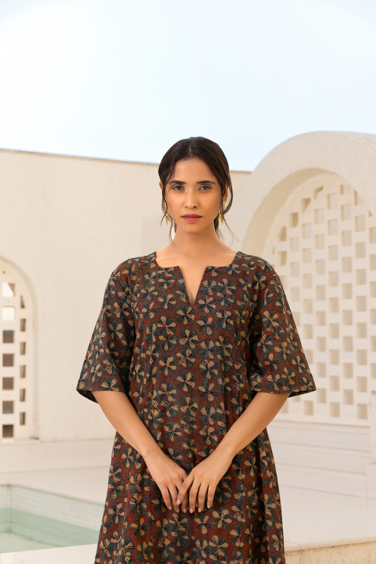 Closeup View of a model showcasing a brown floral printed kurta set with gold accents, paired with striped trousers, styled against an elegant outdoor backdrop.