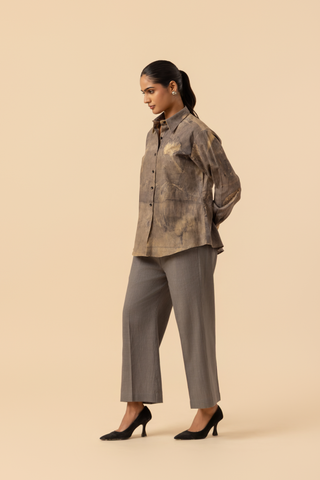 Side view of a model stands against a light beige background, wearing a patterned grey co-ord set which has button-up shirt and wide-legged beige pants, accessorized with heels.
