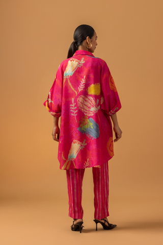 Back view of a model in a vibrant pink tunic with floral patterns co-ord set, paired with striped pink pants and stylish heels, against a warm, neutral background.
