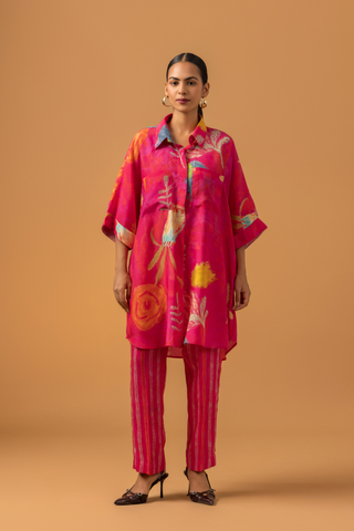 A model in a vibrant pink tunic with floral patterns co-ord set, paired with striped pink pants and stylish heels, against a warm, neutral background.