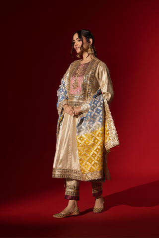 Side view of a Woman in a shimmering gold brocade kurta set stands confidently against a rich red backdrop, showcasing intricate patterns and styling.
