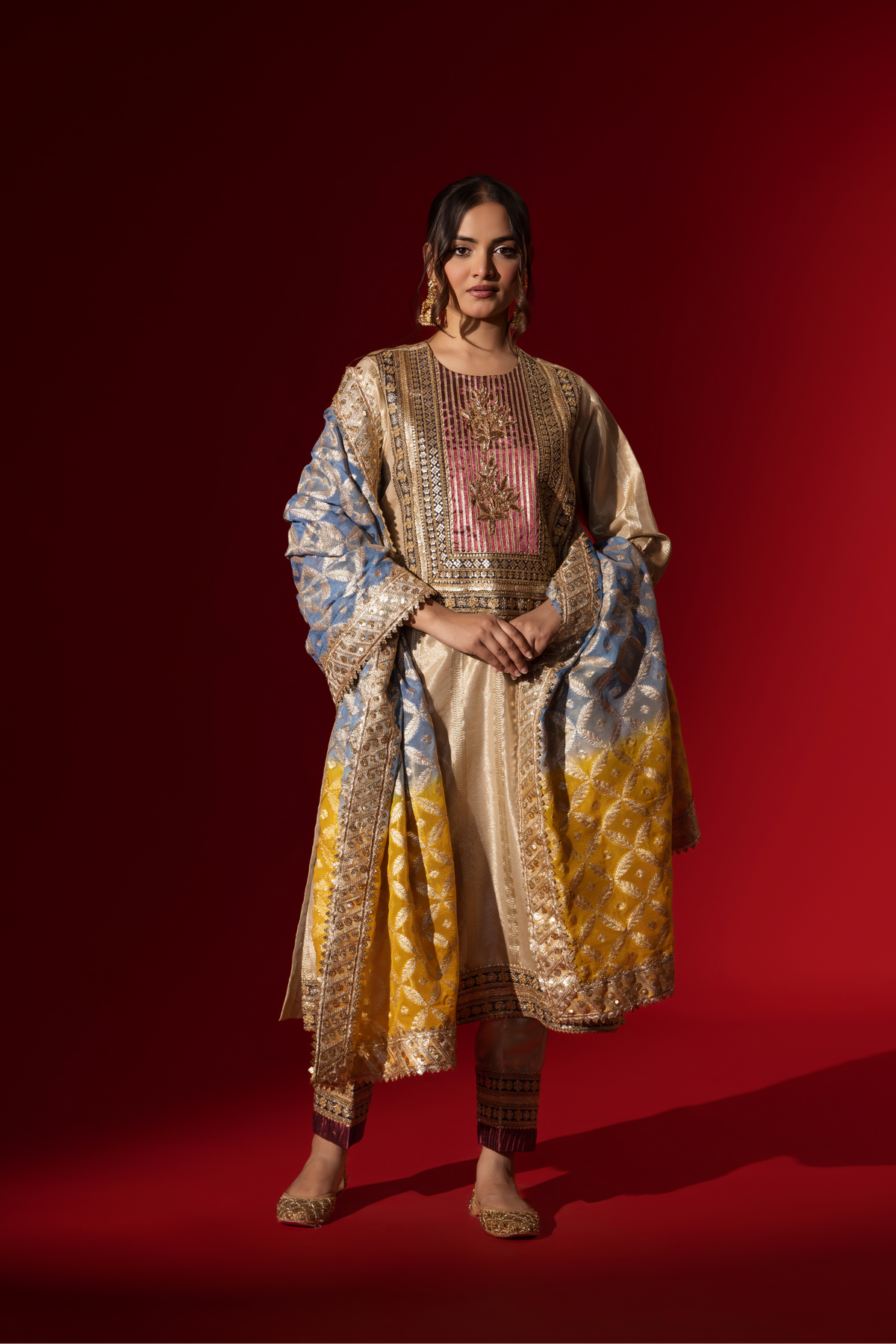 A Woman in a shimmering gold brocade kurta set stands confidently against a rich red backdrop, showcasing intricate patterns and styling.