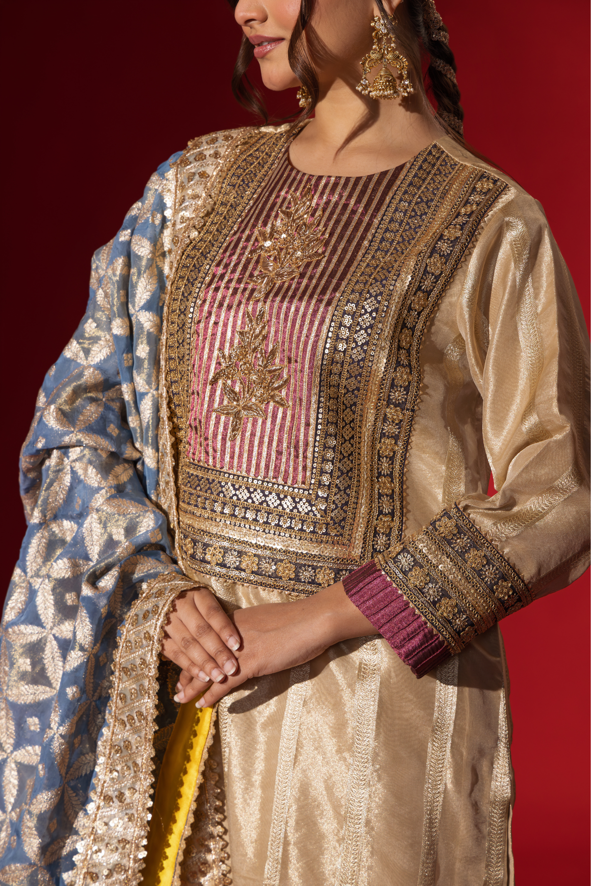 Closeup view of a Woman's hand in a shimmering gold brocade kurta set stands confidently against a rich red backdrop, showcasing intricate patterns and styling.